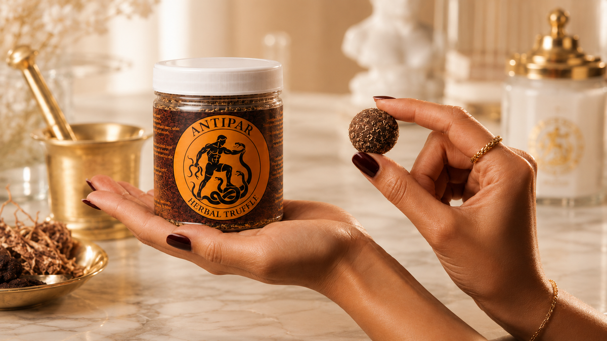 A hand holds a jar of Antipar Herbal Truffles next to another hand holding a single round herbal truffle, set against a warm, softly blurred background of a marble countertop and gold-toned apothecary props.